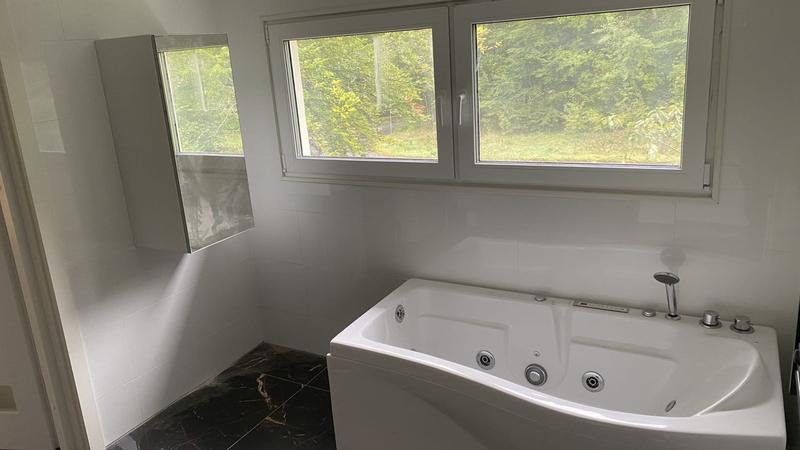 Bubbelbadkar under ett stort fönster i nyrenoverat badrum med vitt kakel och svart klinker